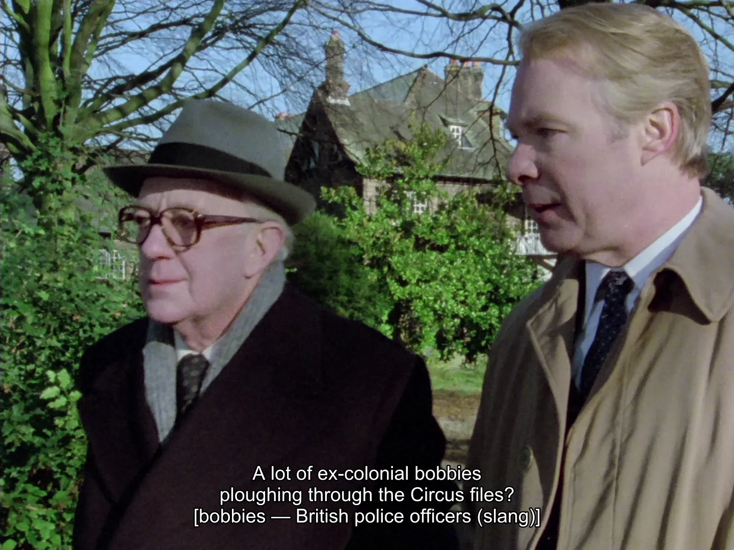 The same frame as the bobbies shot above, now on the English + Explainers track: the dialogue “A lot of ex-colonial bobbies ploughing through the Circus files?” is subtitled with “[bobbies — British police officers (slang)]” in brackets beneath it